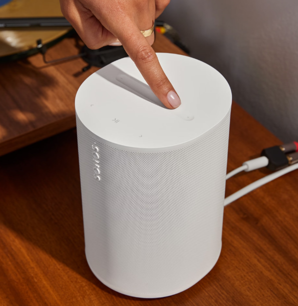 Eine Hand drückt auf die Oberseite eines weißen Sonos-Lautsprechers auf einem Holztisch.