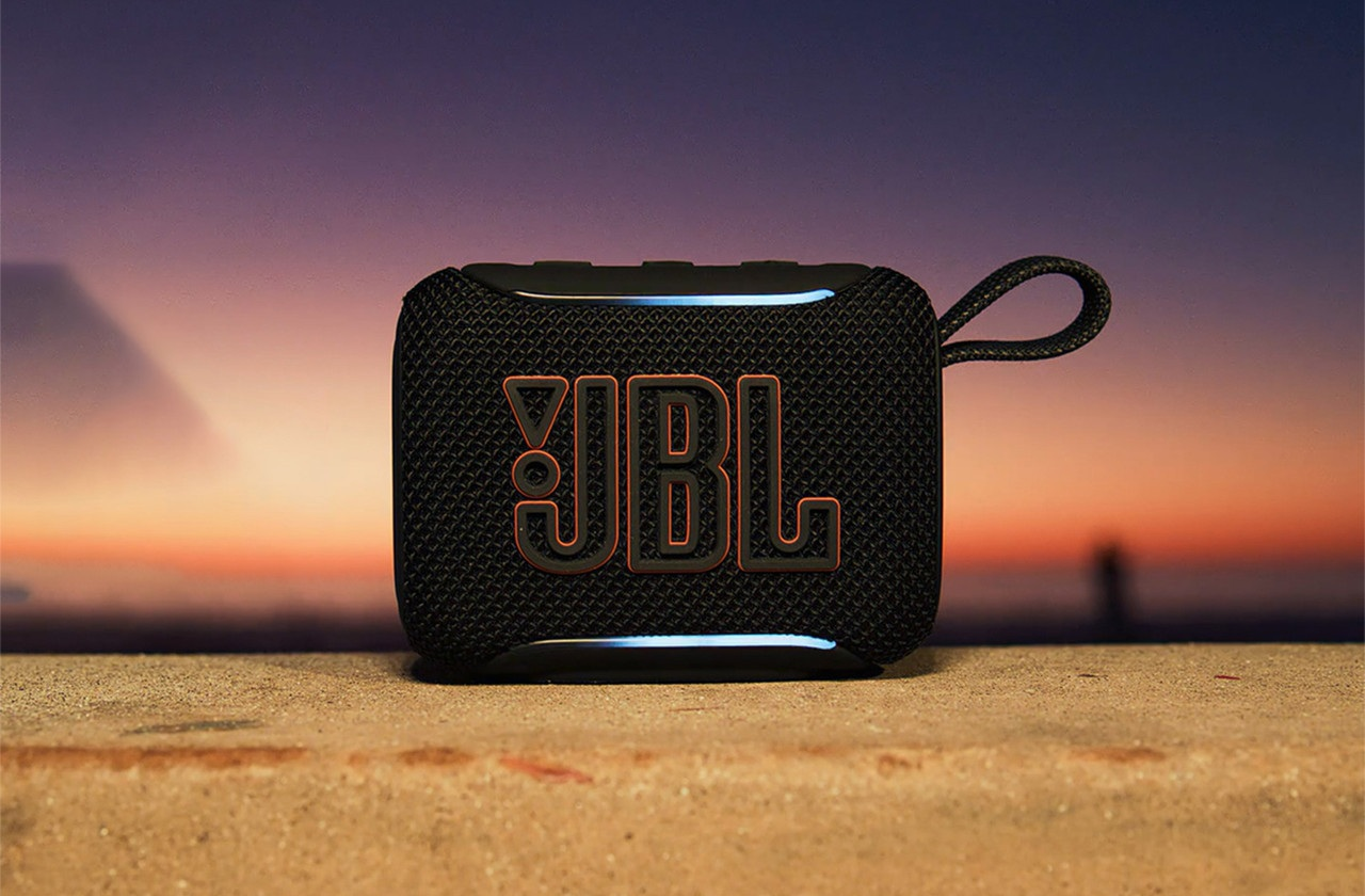 Czarny głośnik JBL z pomarańczowym logo na piaszczystej plaży o zachodzie słońca.