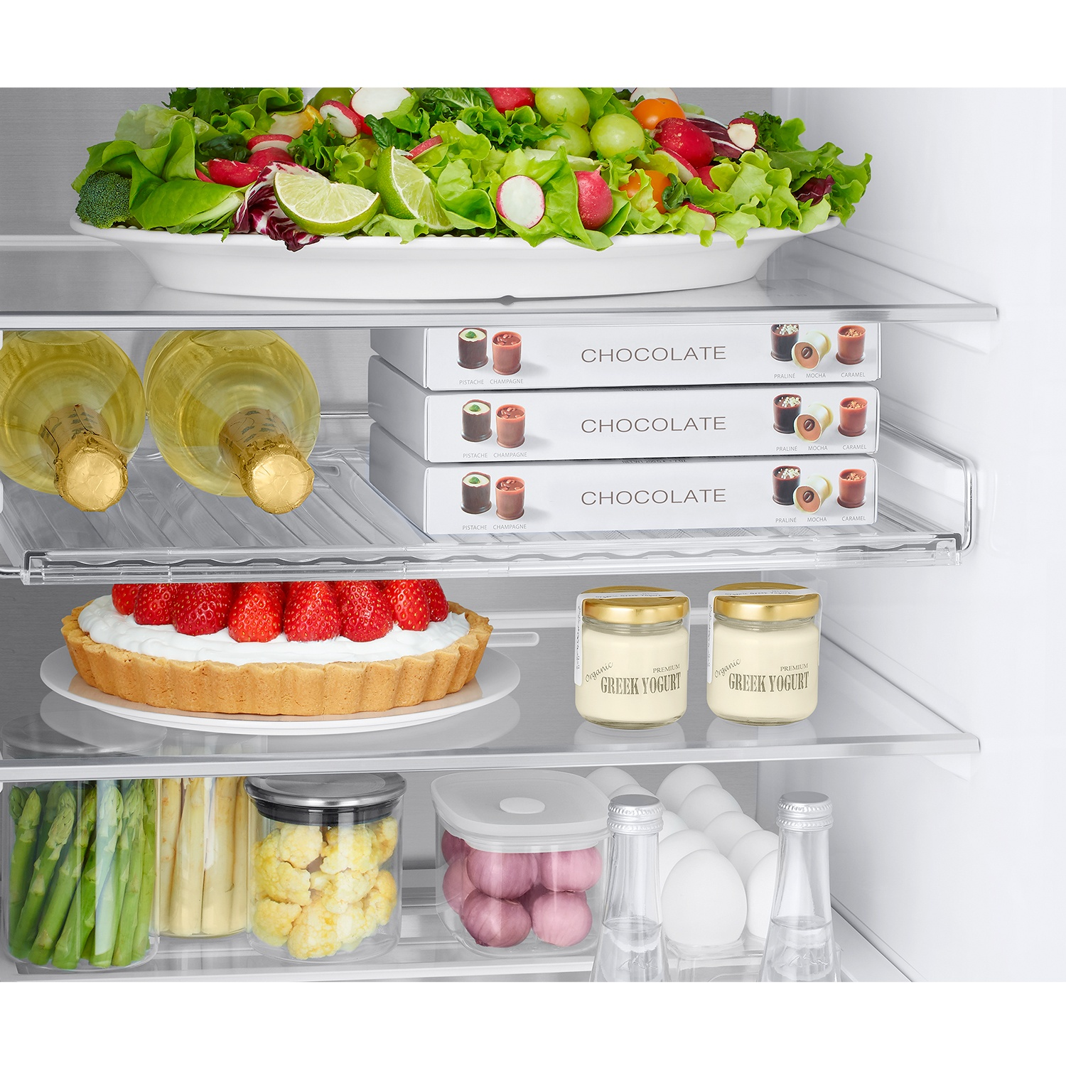 Una balda de refrigerador con ensalada, champán, bombones, tarta de fresa, yogur, espárragos, coliflor y huevos.