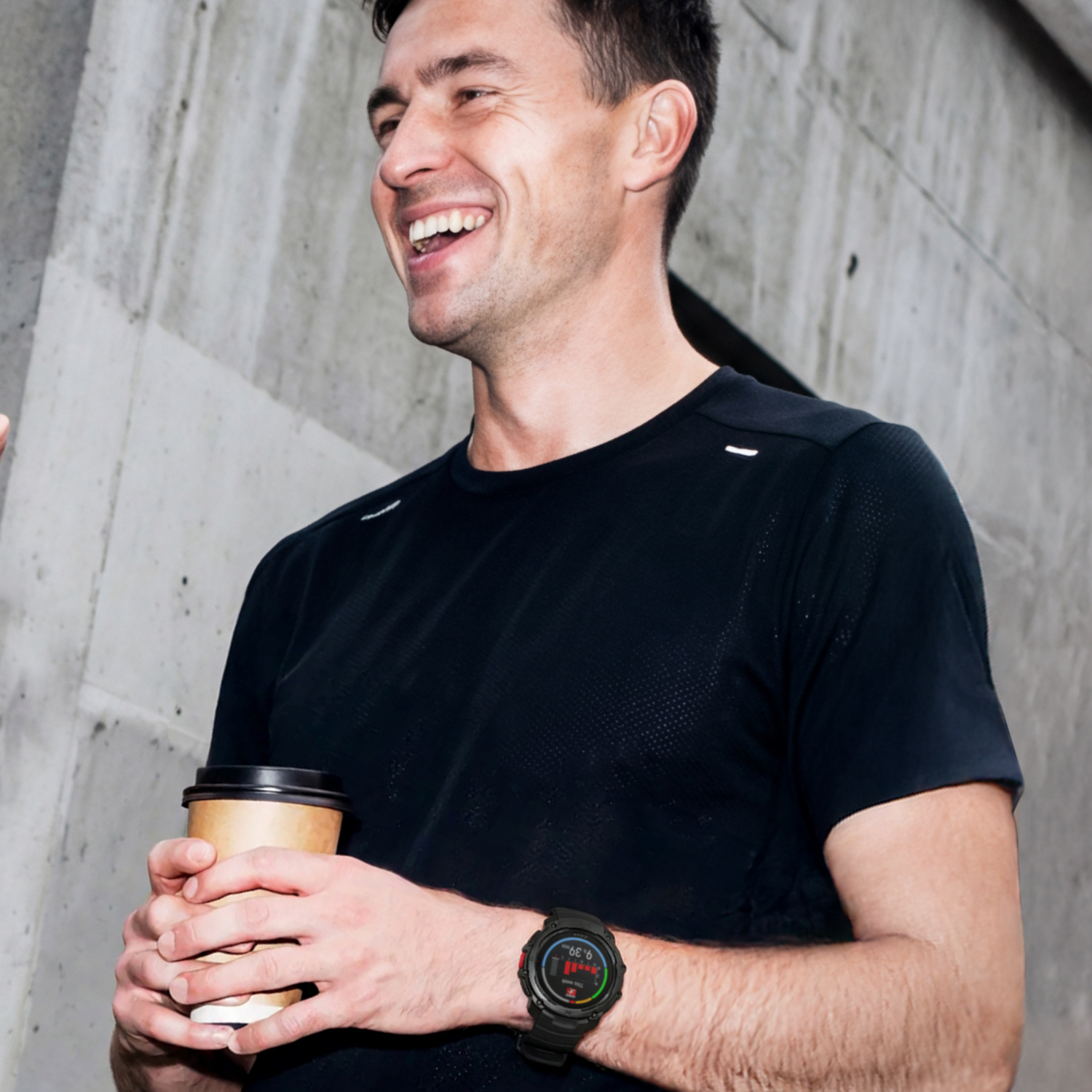 Un hombre sonriente con camiseta negra sostiene un vaso de café de cartón. Lleva un reloj inteligente.