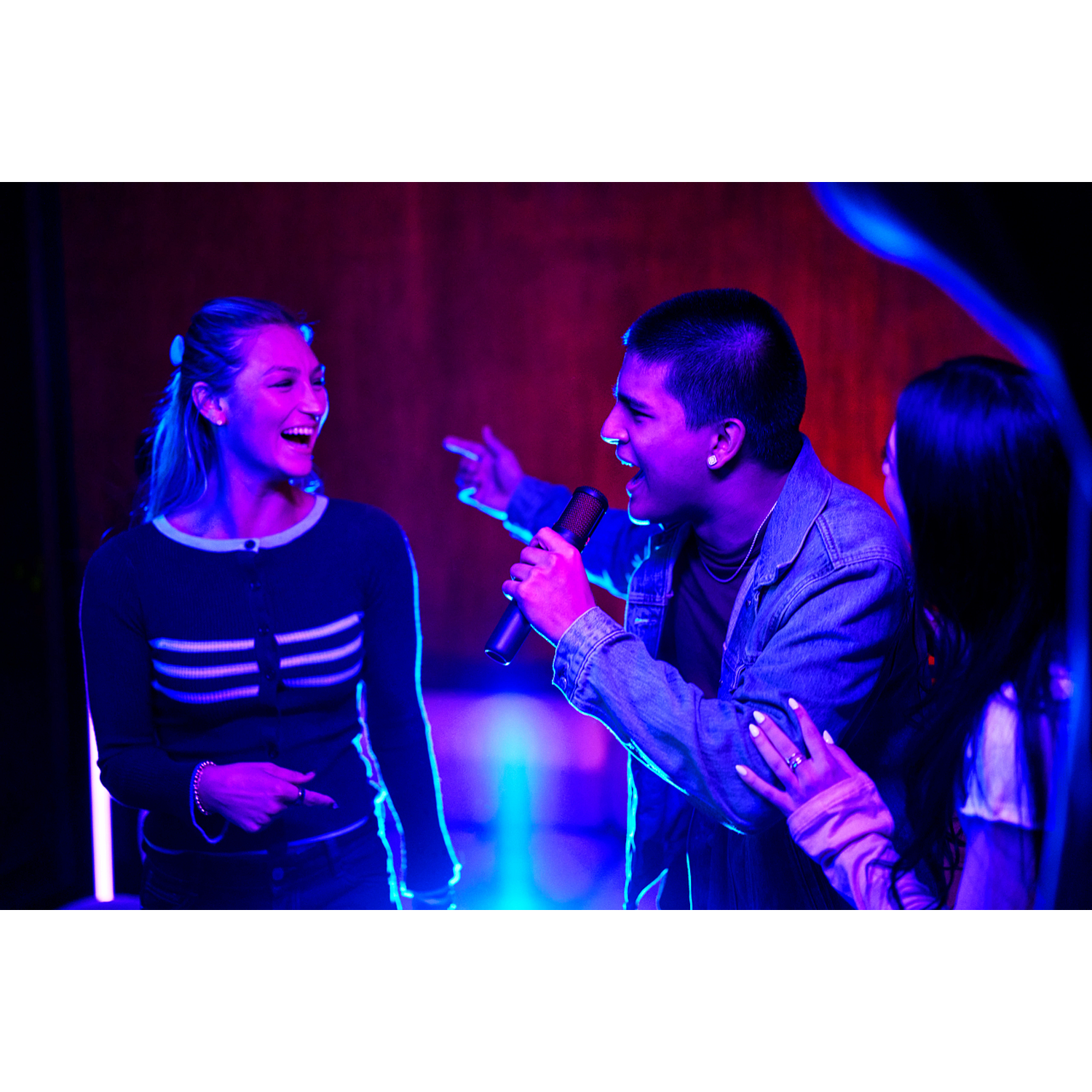Three young people sing in a room with blue and red lights. One sings into a microphone.