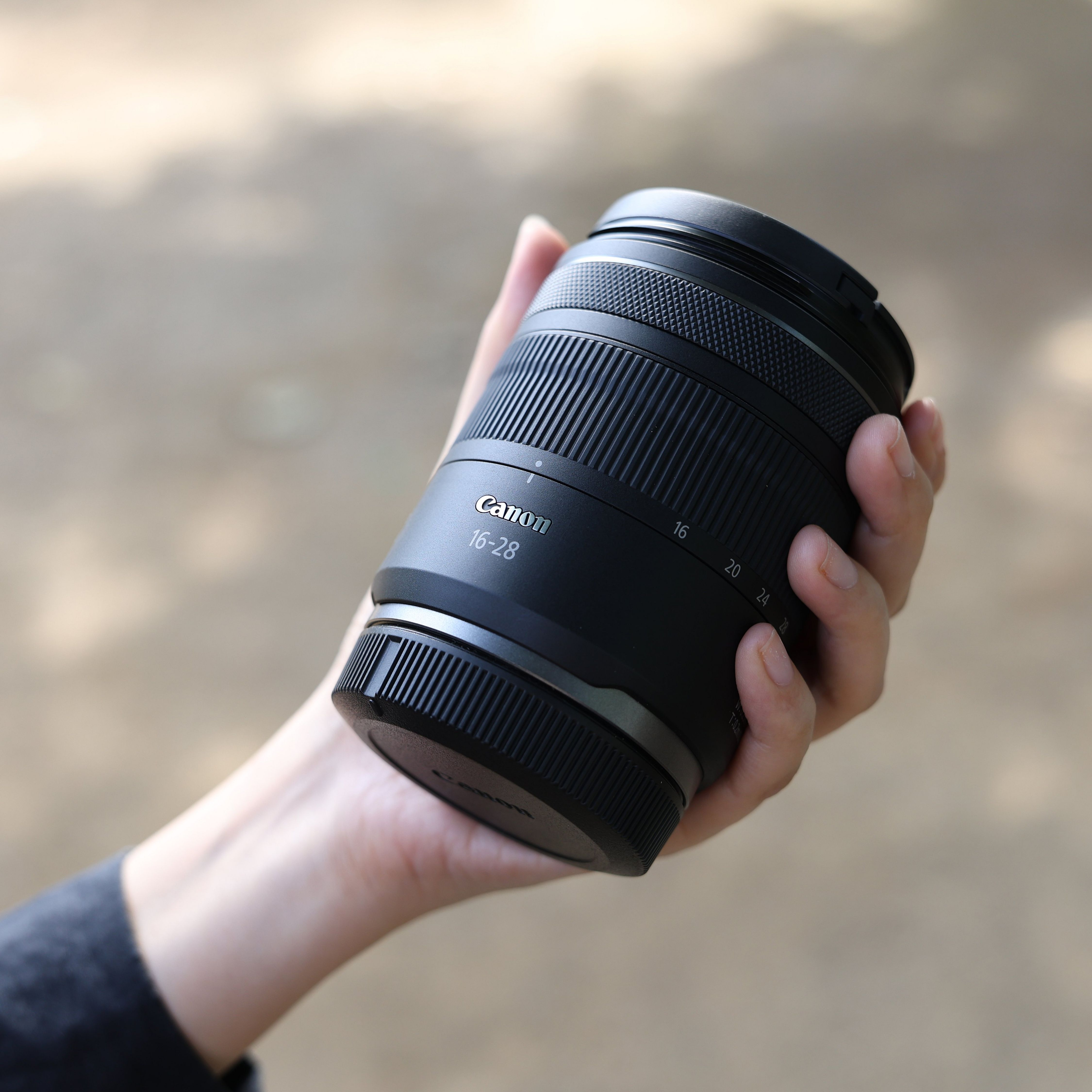 Une main tient un objectif Canon noir 16-28mm sur fond flou.