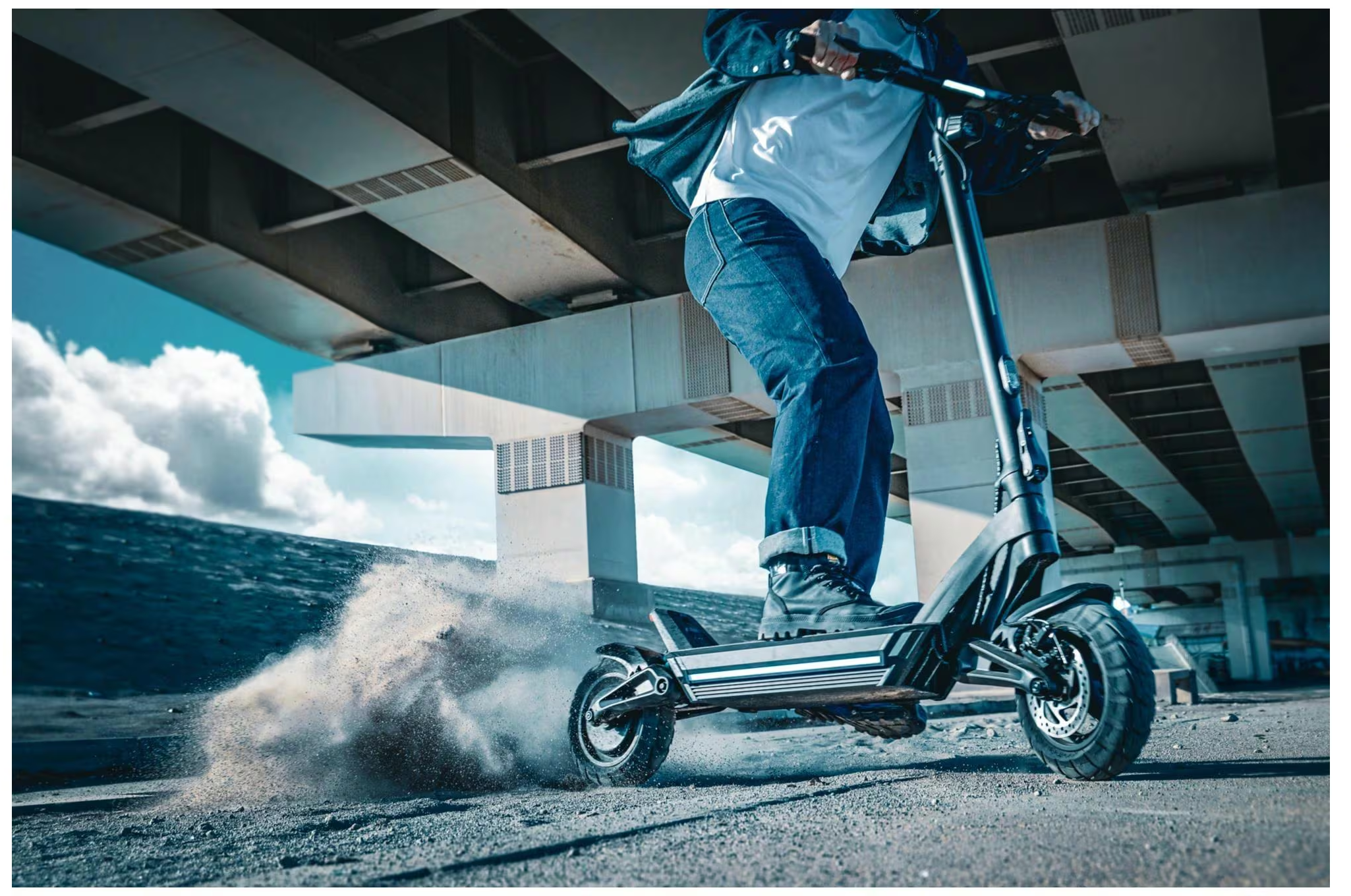 Une personne conduit un scooter électrique, soulevant de la poussière, sous un pont.
