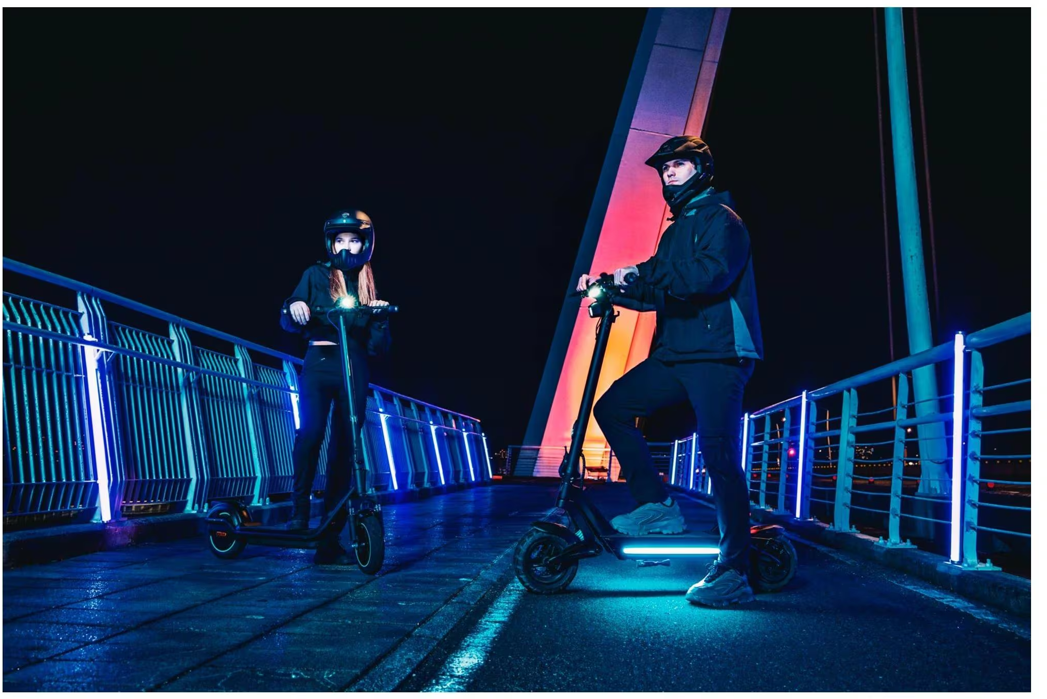 Deux personnes avec des trottinettes électriques sur un pont la nuit, éclairées par des bandes LED bleues.