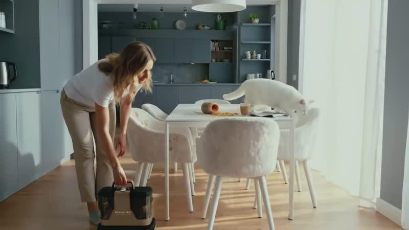 Eine Frau mit einem Staubsauger in einer Küche, eine weiße Katze auf dem Tisch.