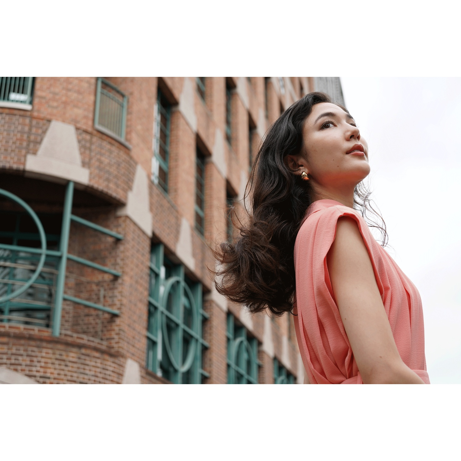 A woman in a coral dress stands in front of a brick building.