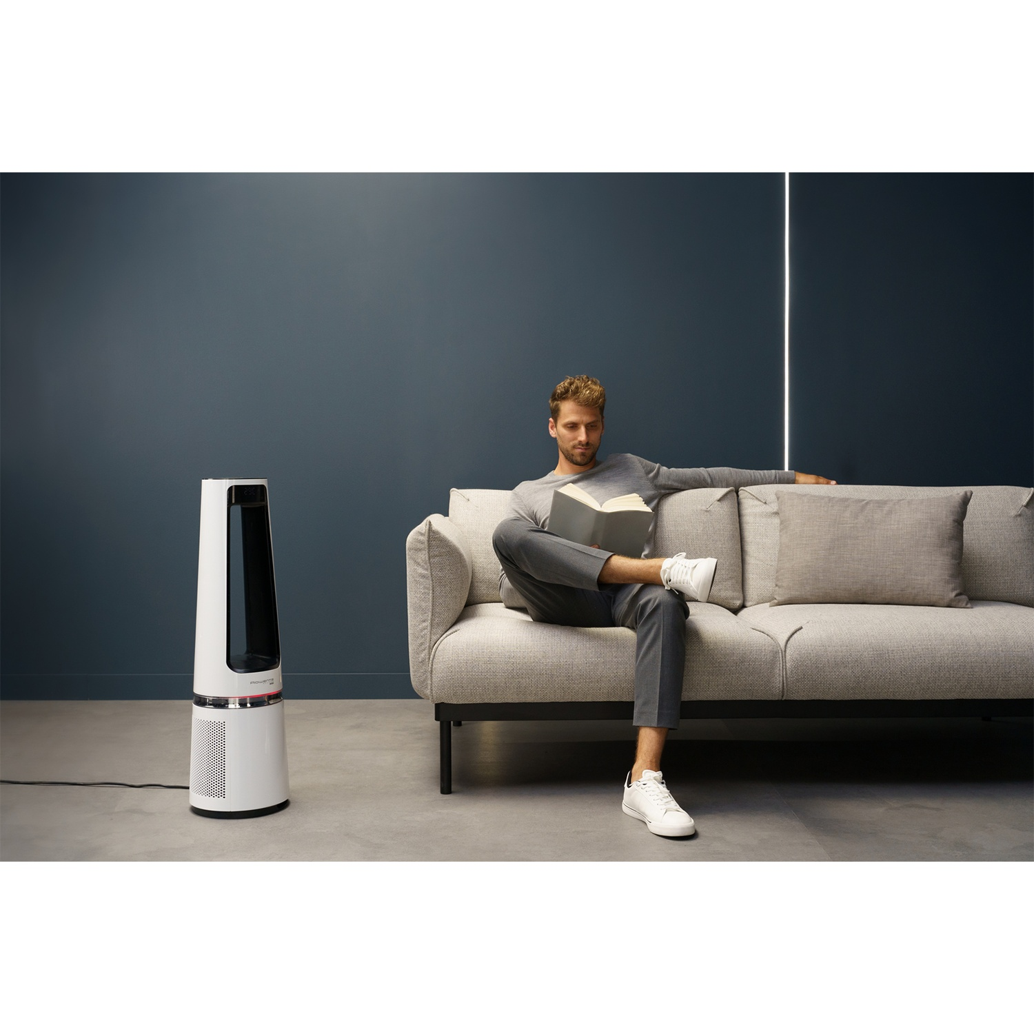 Man reading a book on a sofa next to a white tower fan.