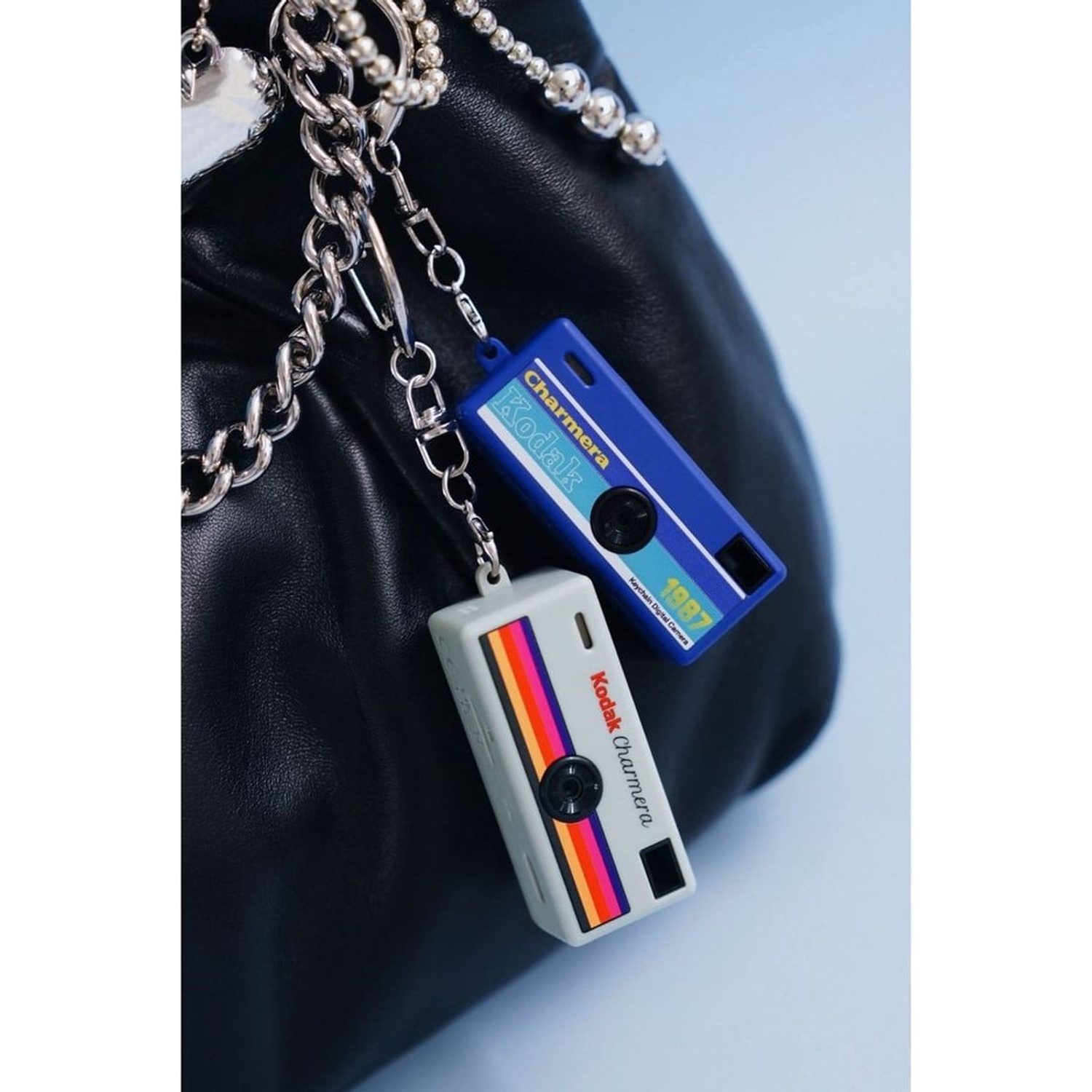 Two Kodak Charmera keychain cameras, one blue, one gray, on a black bag.