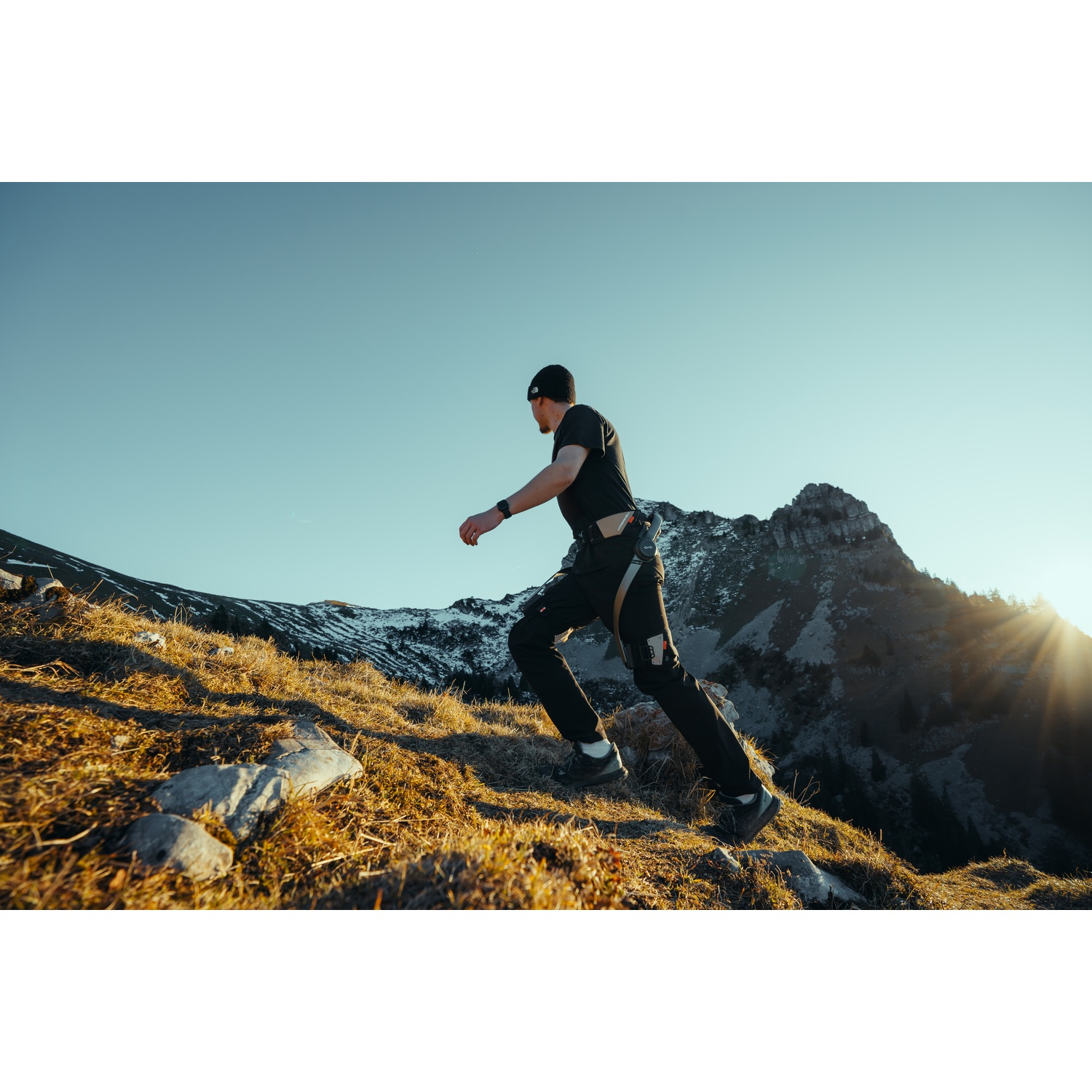 Uomo corre in salita su un pendio montano erboso con sole e vetta rocciosa.