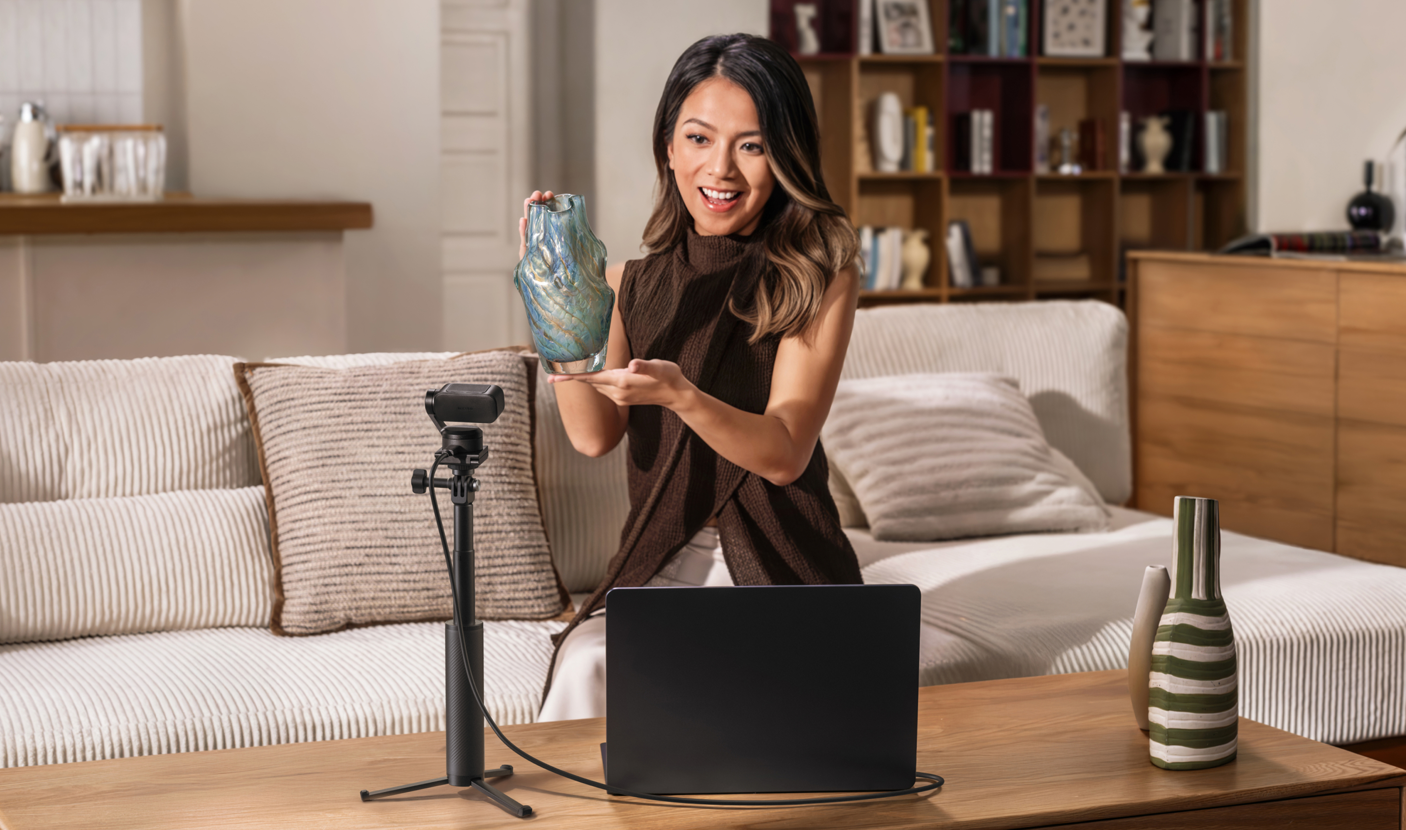 Eine Frau hält eine blaue Vase, ein Laptop und eine Kamera stehen auf einem Tisch.