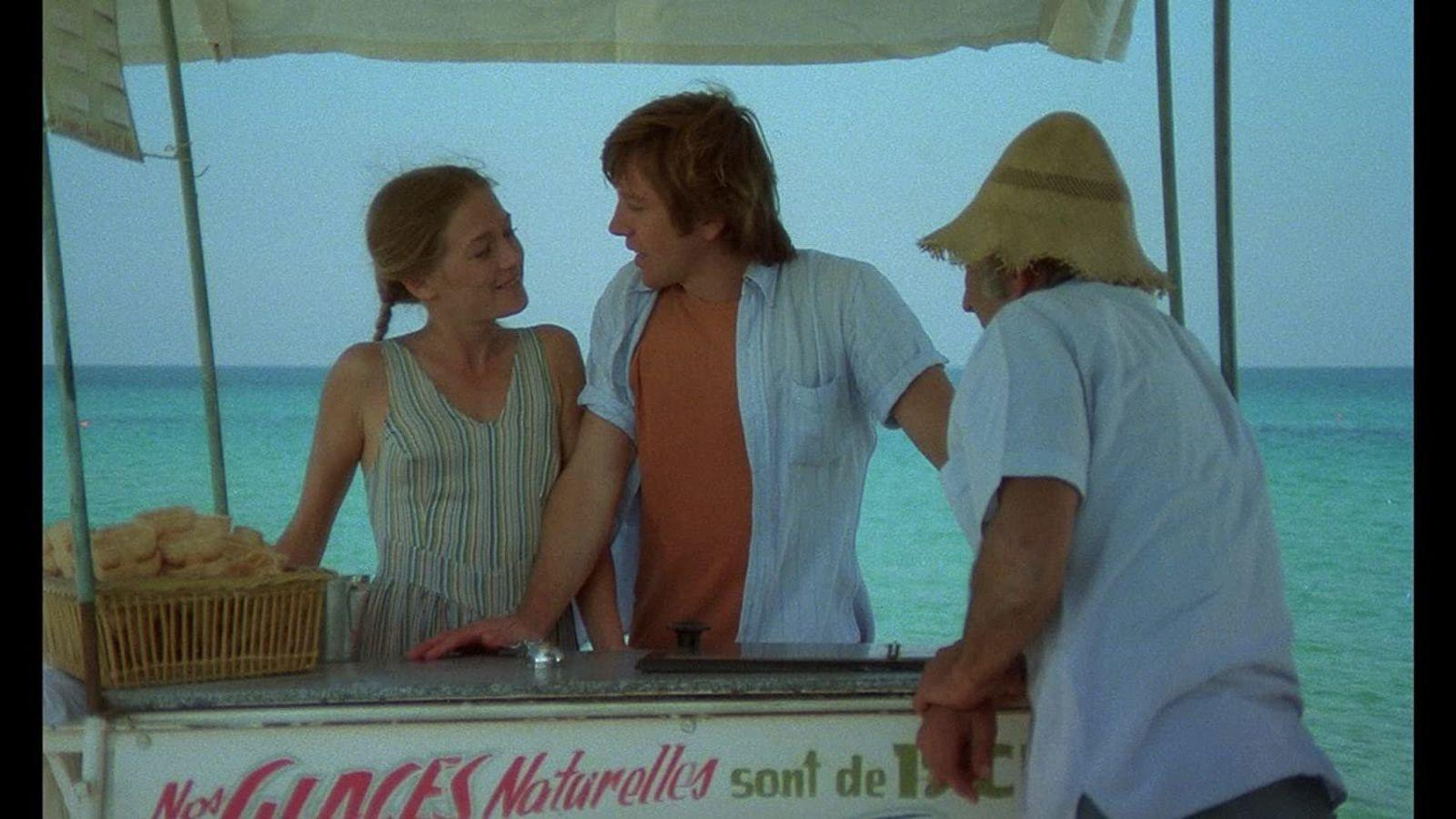 Drei Personen an einem Eisstand am Meer. Eine Frau und ein Mann schauen sich an.