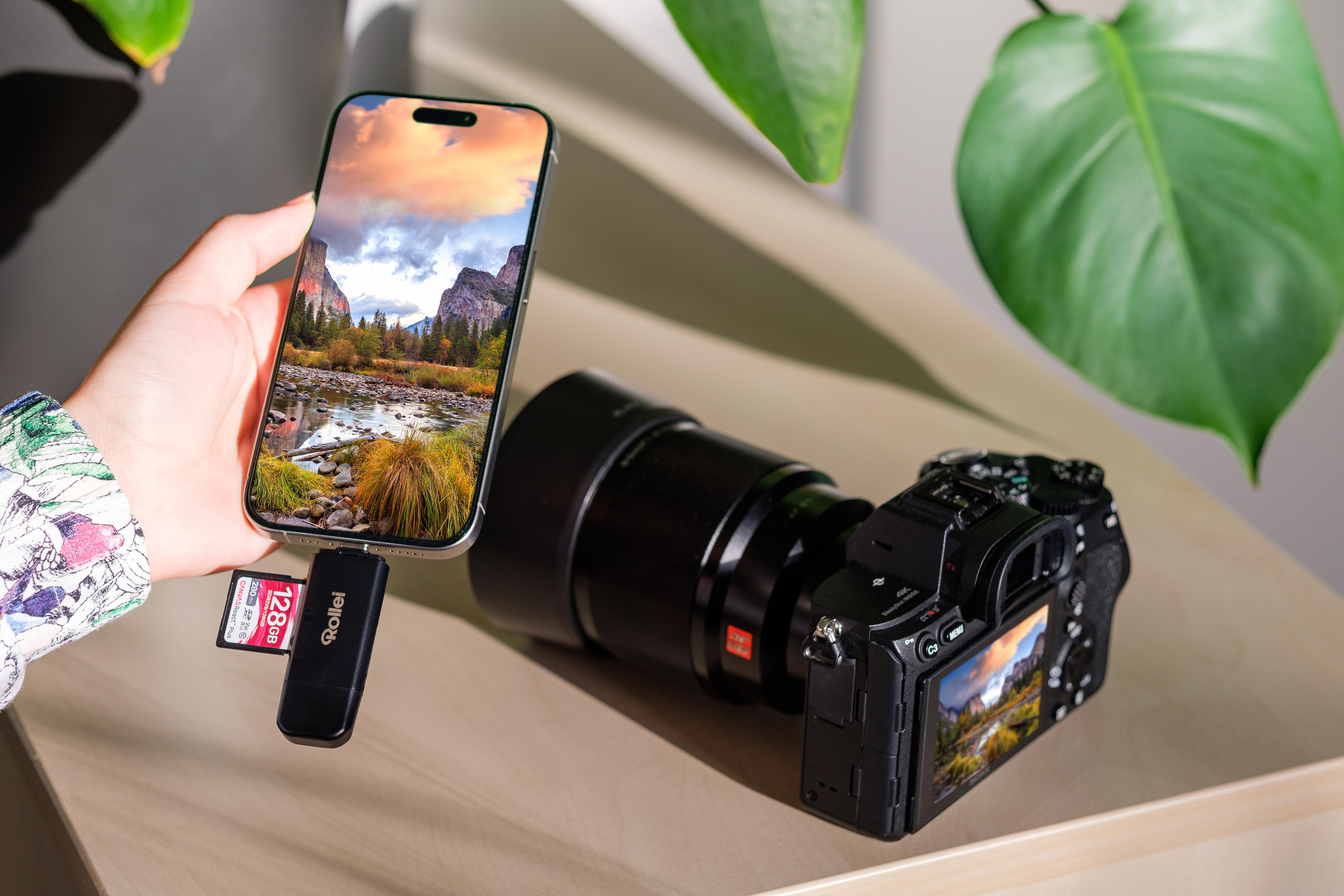 Eine Hand hält ein Smartphone mit Berglandschaft. daneben eine Kamera und ein Speicherkartenleser.