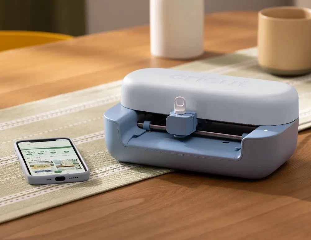 Una máquina cortadora Cricut azul y blanca está al lado de un smartphone sobre una mesa.