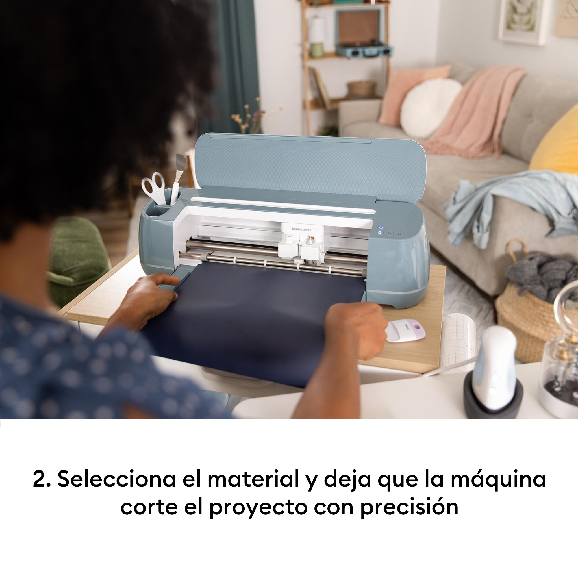 Una persona coloca material azul oscuro en una máquina de corte azul. Sala de estar al fondo.