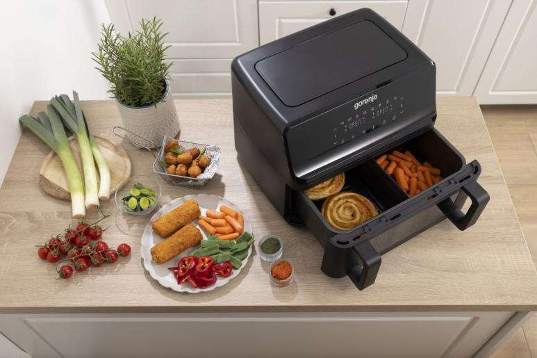 Fekete légkeveréses fritőz nyitott fiókkal, amelyben tekercsek és répa látható. Hozzávalók a közelben.