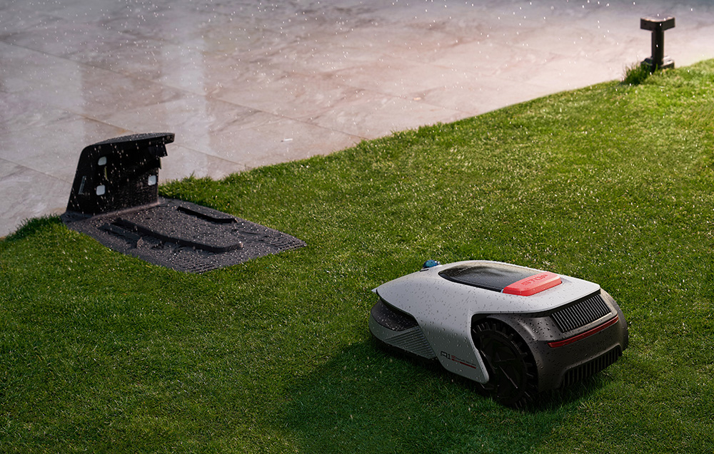 Biała kosiarka robotyczna z czerwonym przyciskiem stop na zielonej trawie w deszczu.