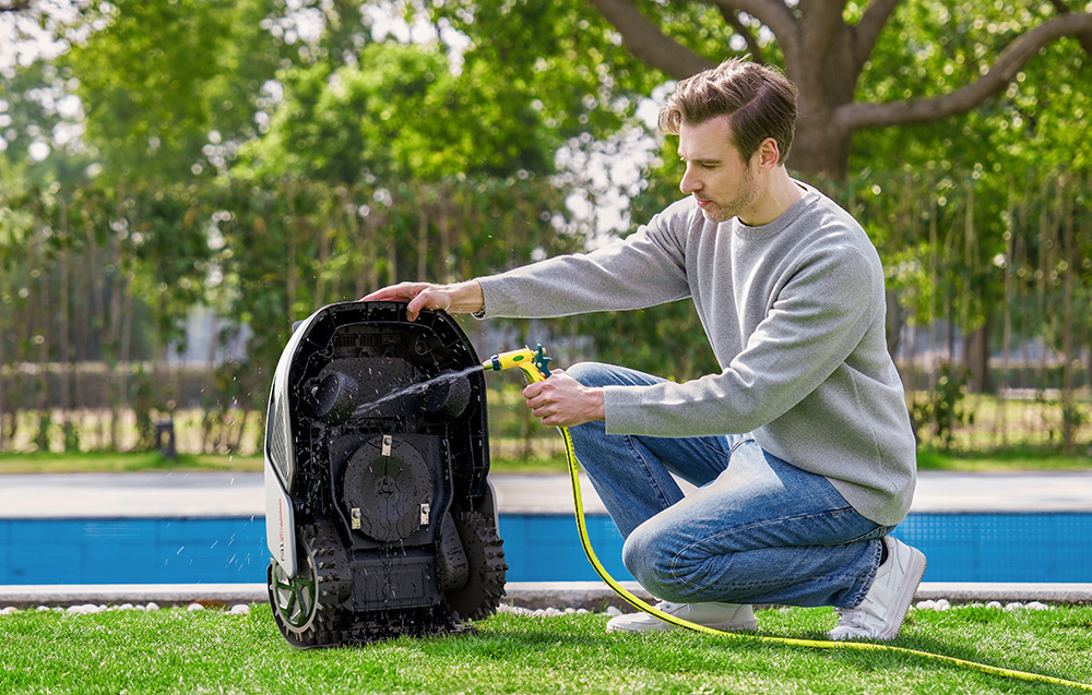 Mężczyzna myje robota koszącego żółtym wężem w ogrodzie z basenem.