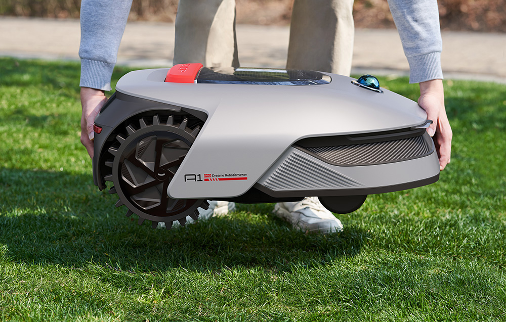 Mężczyzna podnosi szarą kosiarkę automatyczną z dużymi kołami na zielonym trawniku.