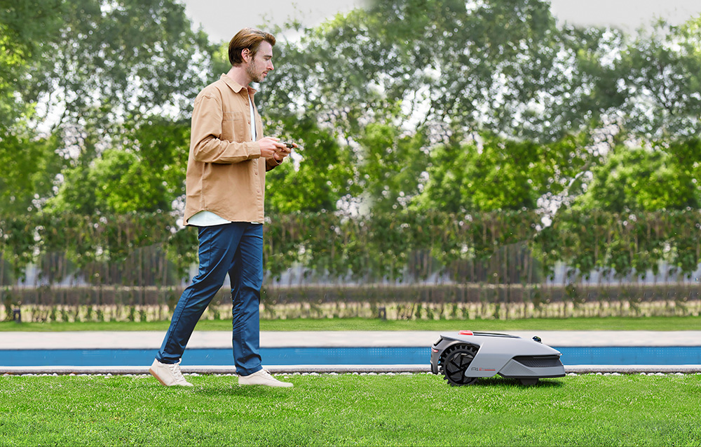 Mężczyzna z pilotem steruje robotem koszącym na zielonej trawie przy niebieskim basenie.