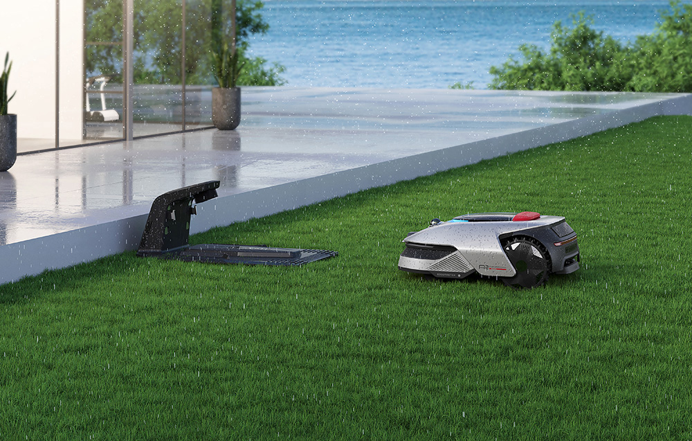 Srebrny robot koszący na zielonym trawniku przed tarasem, w tle morze.