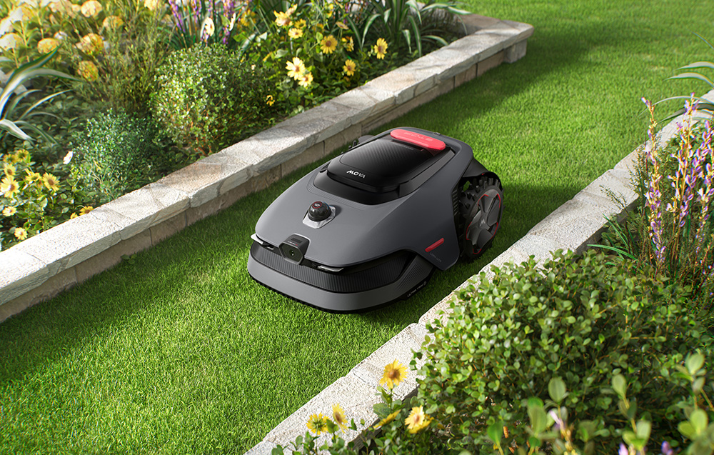 Szara kosiarka automatyczna z czerwonym akcentem na zielonym trawniku, otoczona kwiatami i krzewami.