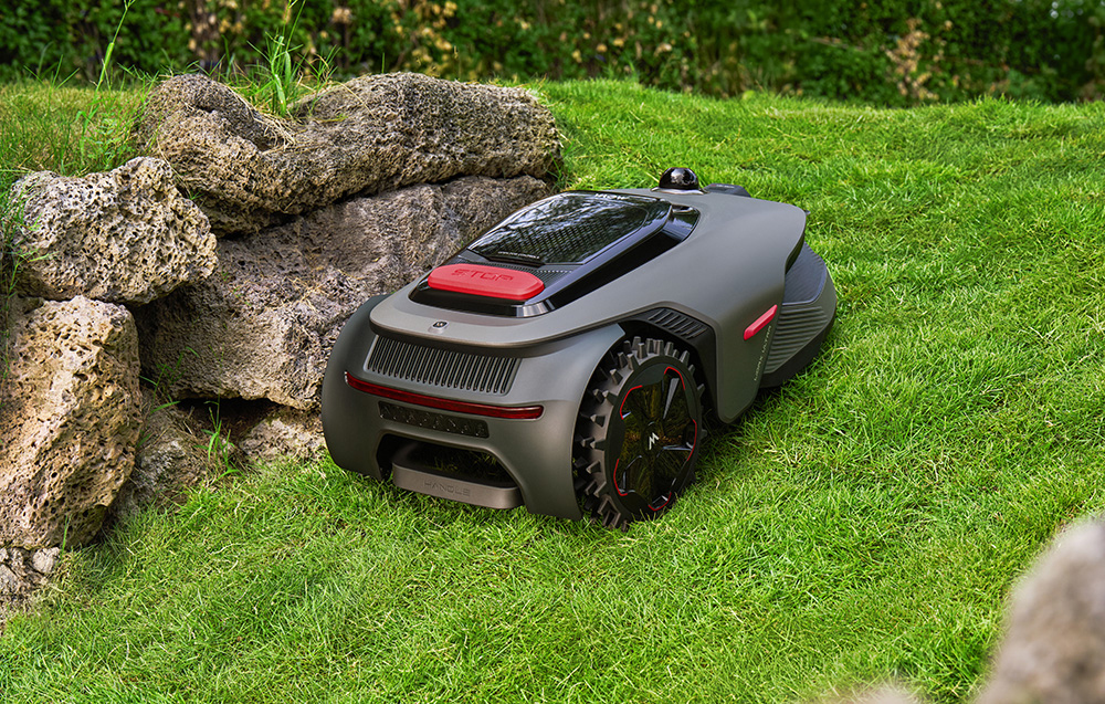 Szara kosiarka automatyczna z czerwonymi akcentami stoi na zielonym trawniku obok skał.