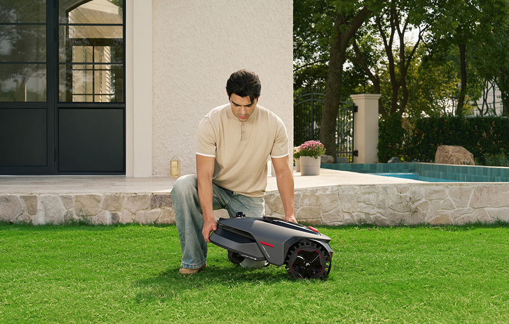 Mężczyzna klęczy na trawie, trzymając robota koszącego. W tle dom i basen.