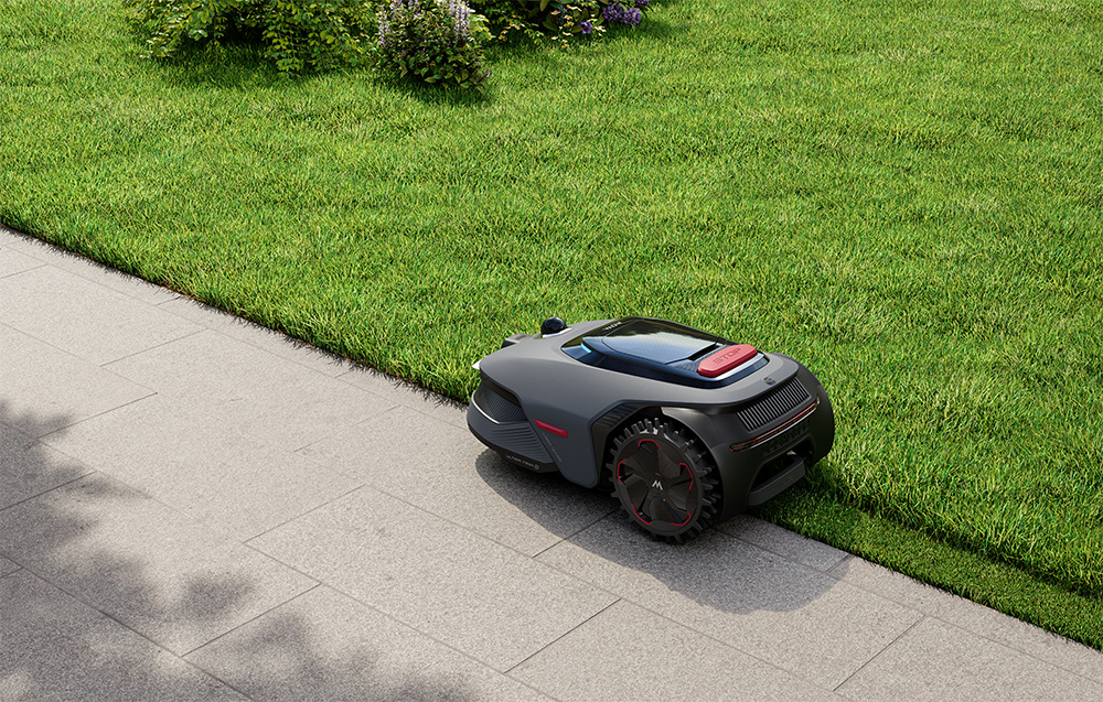 Szara kosiarka automatyczna z czerwonymi akcentami stoi na brukowanej ścieżce obok trawnika.