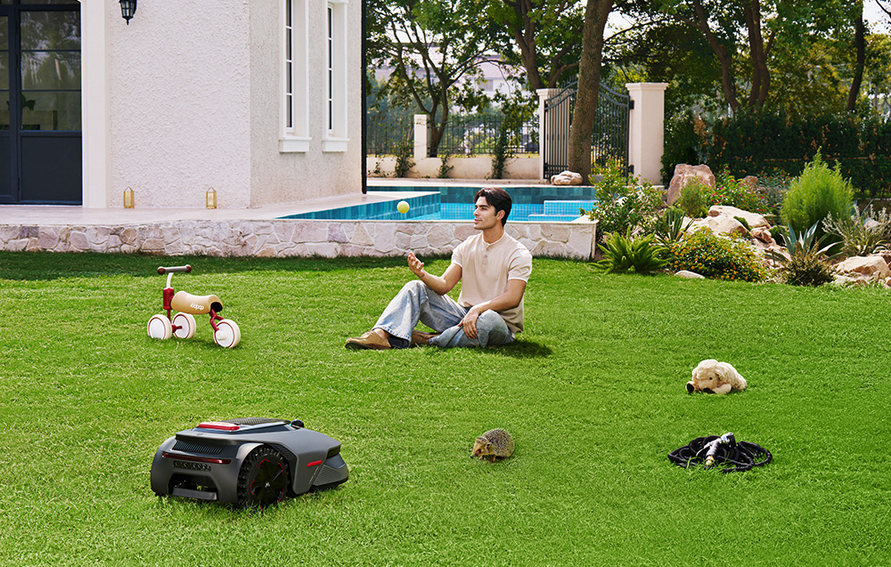 Mężczyzna rzuca piłkę w ogrodzie z robotem koszącym, jeżem, rowerkiem i psem.