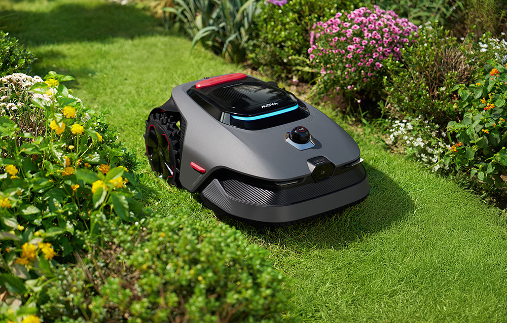 Szara kosiarka automatyczna stoi na zielonej trawie obok kolorowych kwiatów.