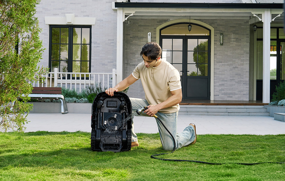 Mężczyzna klęczy na trawie, czyszcząc robota koszącego wężem.