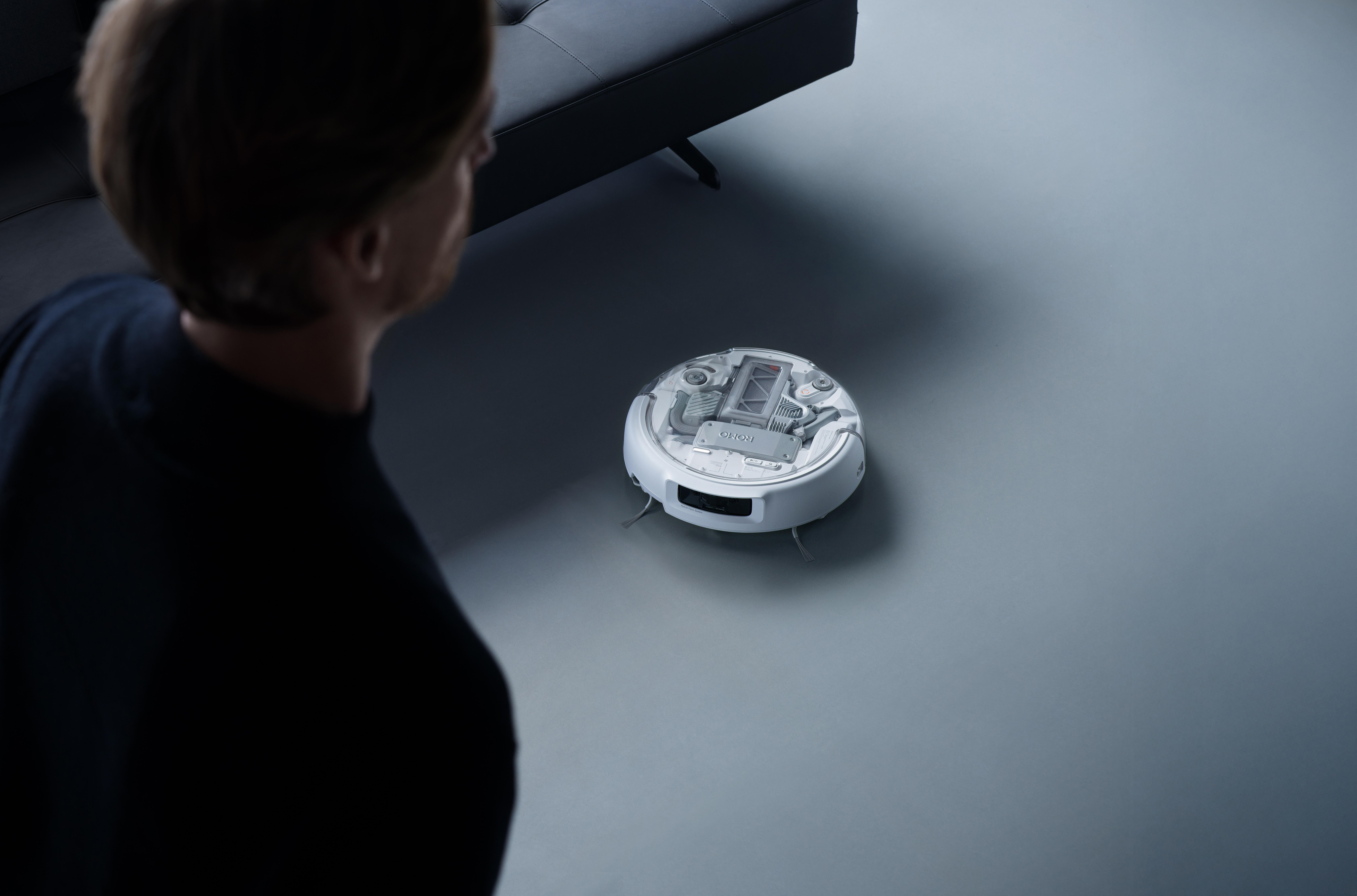 Mann blickt auf weißen Saugroboter neben dunklem Sofa auf grauem Boden.