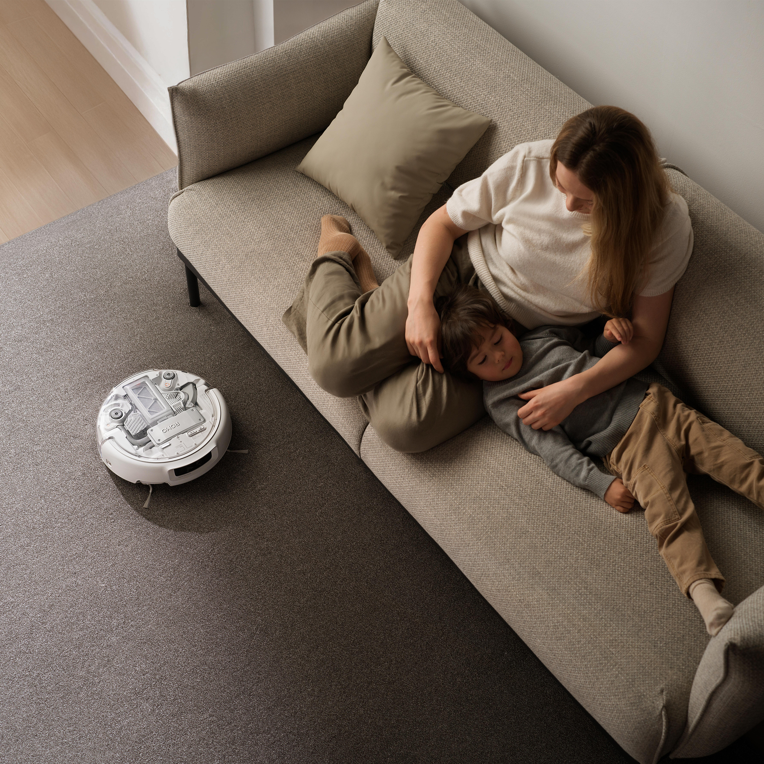 Ein Roboterstaubsauger reinigt den Teppich neben einer Couch, auf der eine Frau und ein Kind liegen.