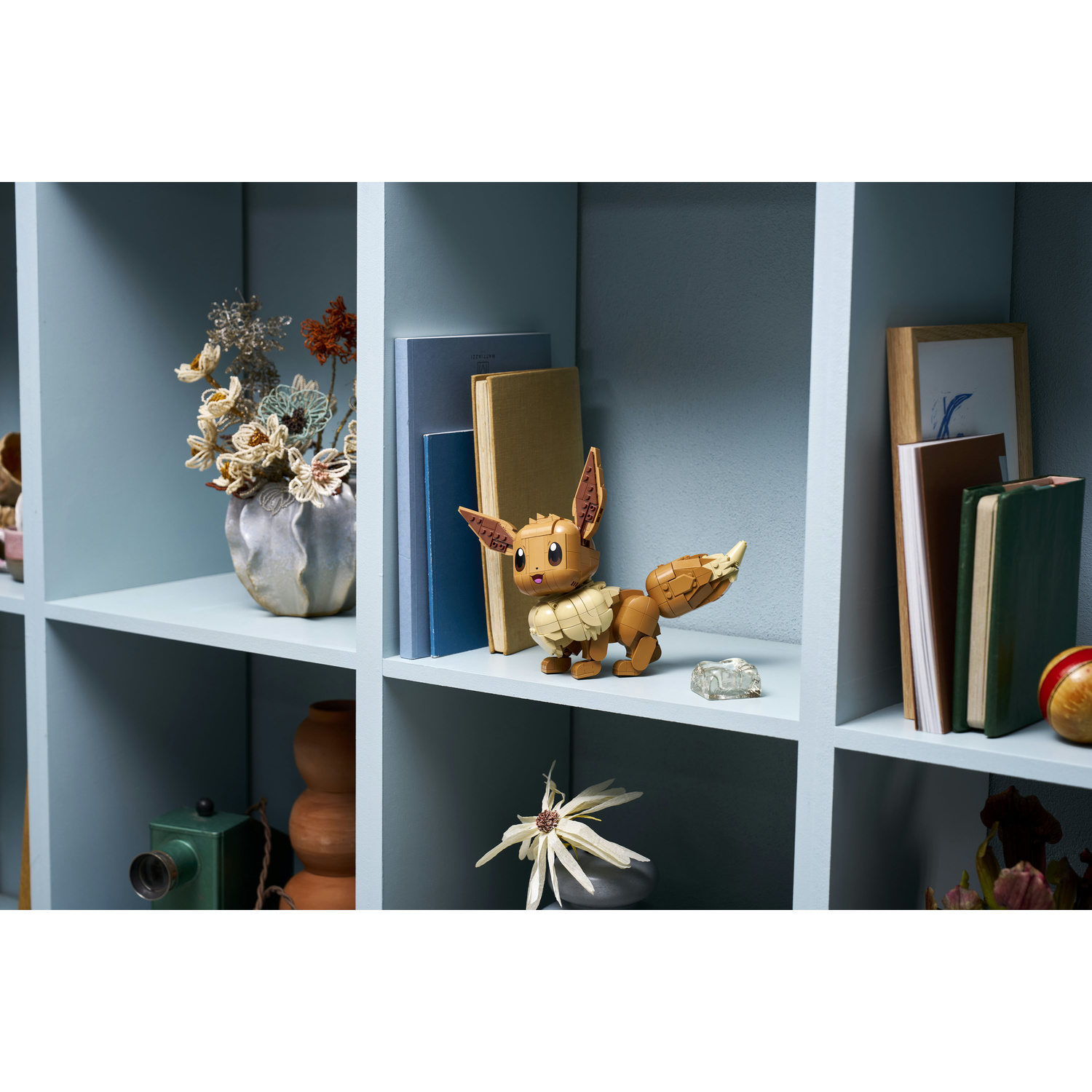 Un giocattolo Eevee si trova su uno scaffale accanto a libri e un vaso di fiori.