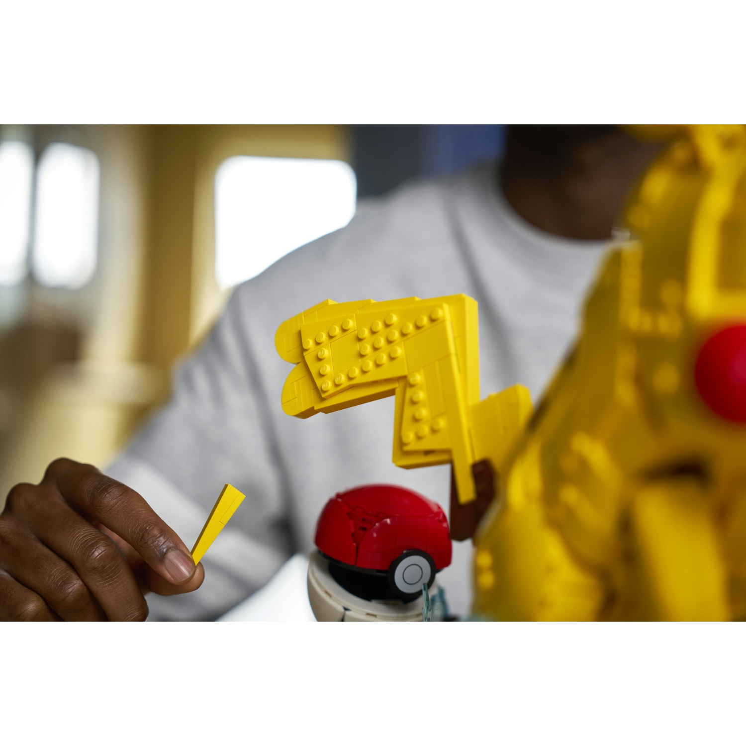 Una mano tiene un mattoncino Lego giallo sopra una Poké Ball e strutture Lego gialle.