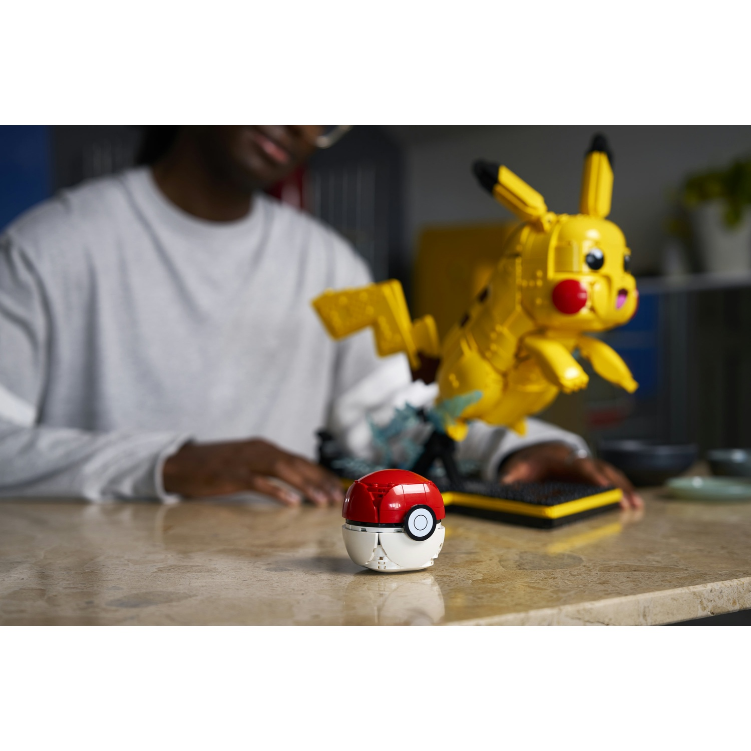 Una persona seduta a un tavolo con un modello di Pokeball e una costruzione Pikachu gialla davanti.