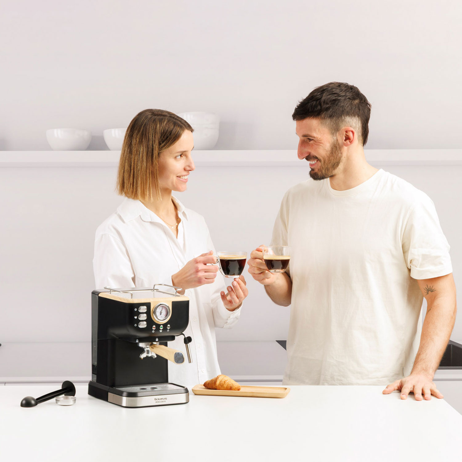 Hombre y mujer brindando con café, máquina de espresso y croissant en la encimera.