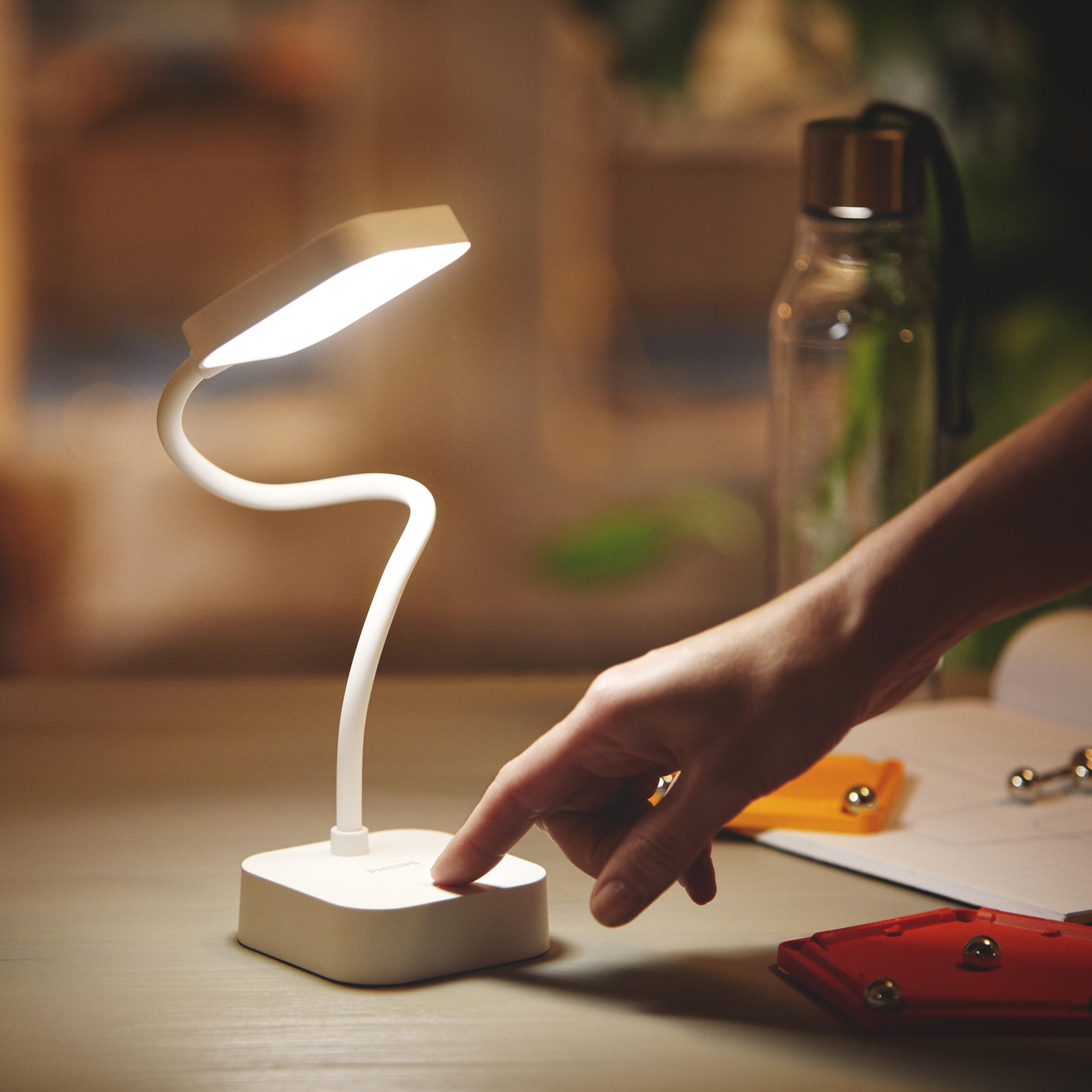 Eine Hand schaltet eine weiße Schreibtischlampe ein. Auf dem Tisch liegen Objekte.