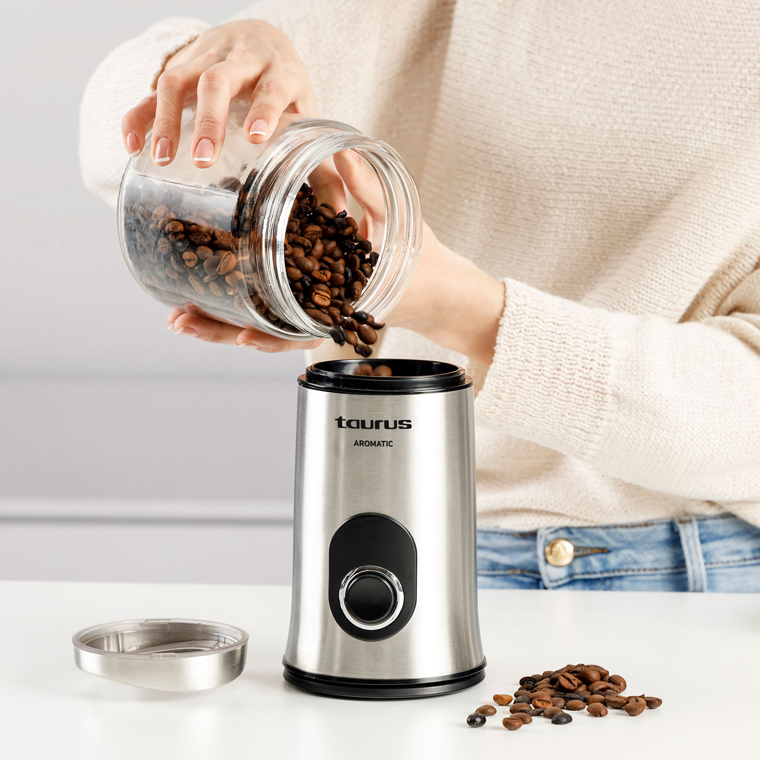 Una persona vierte granos de café de un frasco en un molinillo plateado.