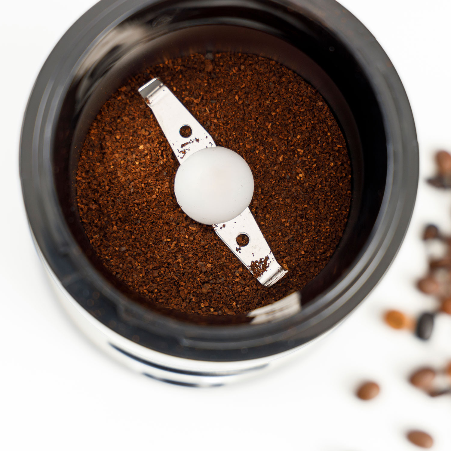 Molinillo de café con café molido y cuchillas. Hay granos de café esparcidos cerca.