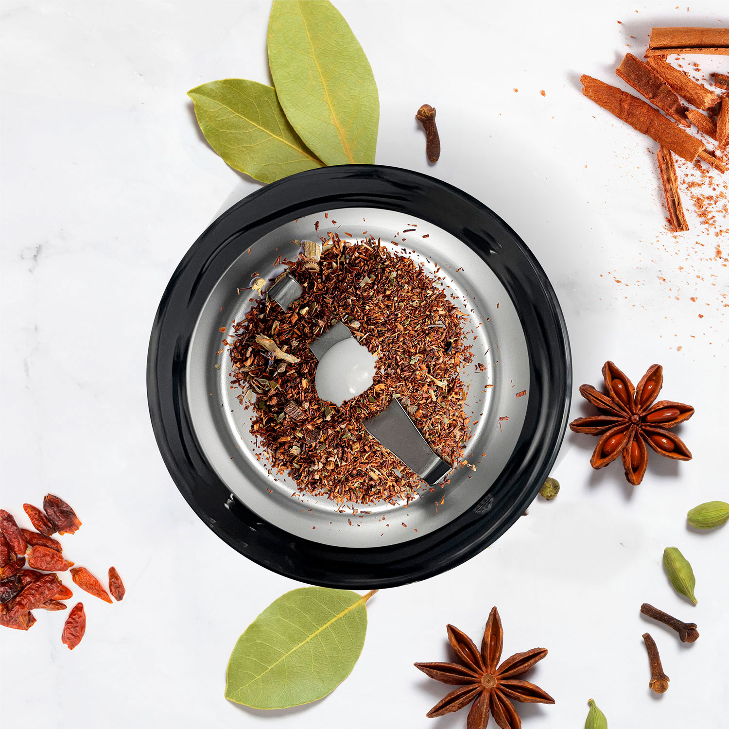 Un molinillo de especias con té molido, rodeado de anís estrellado, canela, clavo, cardamomo, hojas de laurel y chiles.