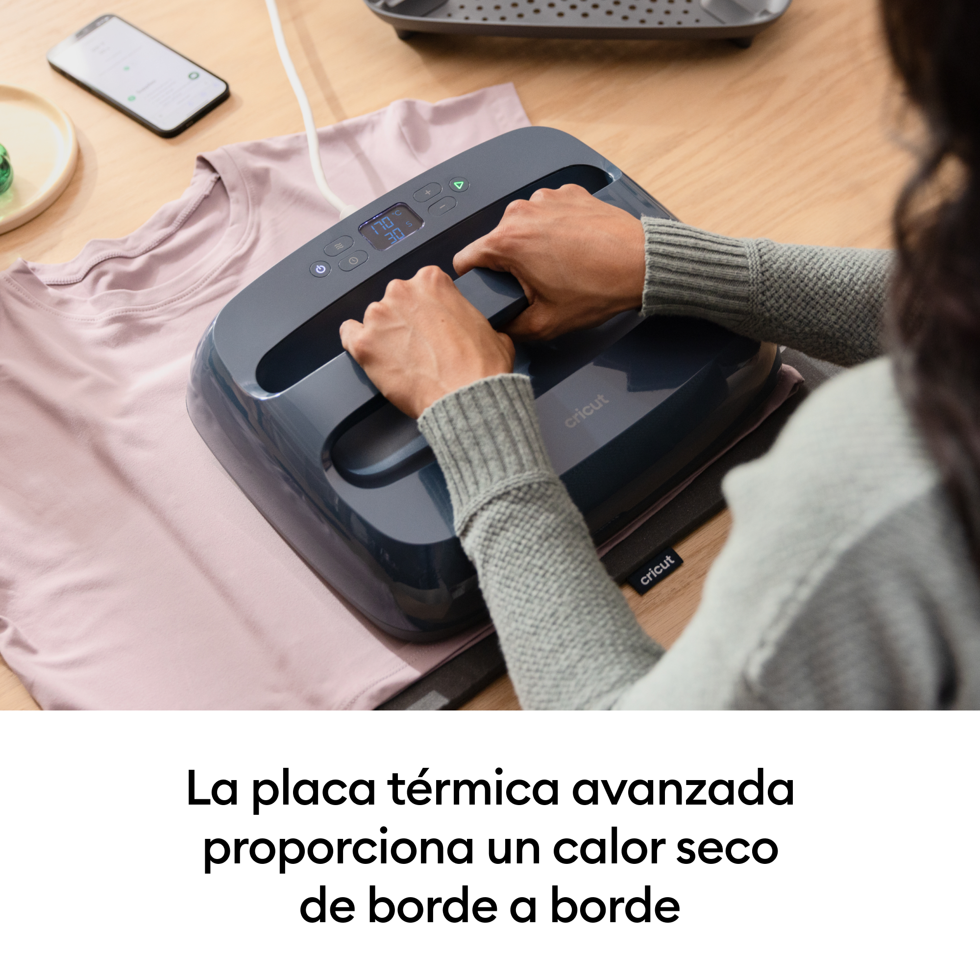 Una persona usa una plancha térmica en una camiseta rosa. Un teléfono está cerca.