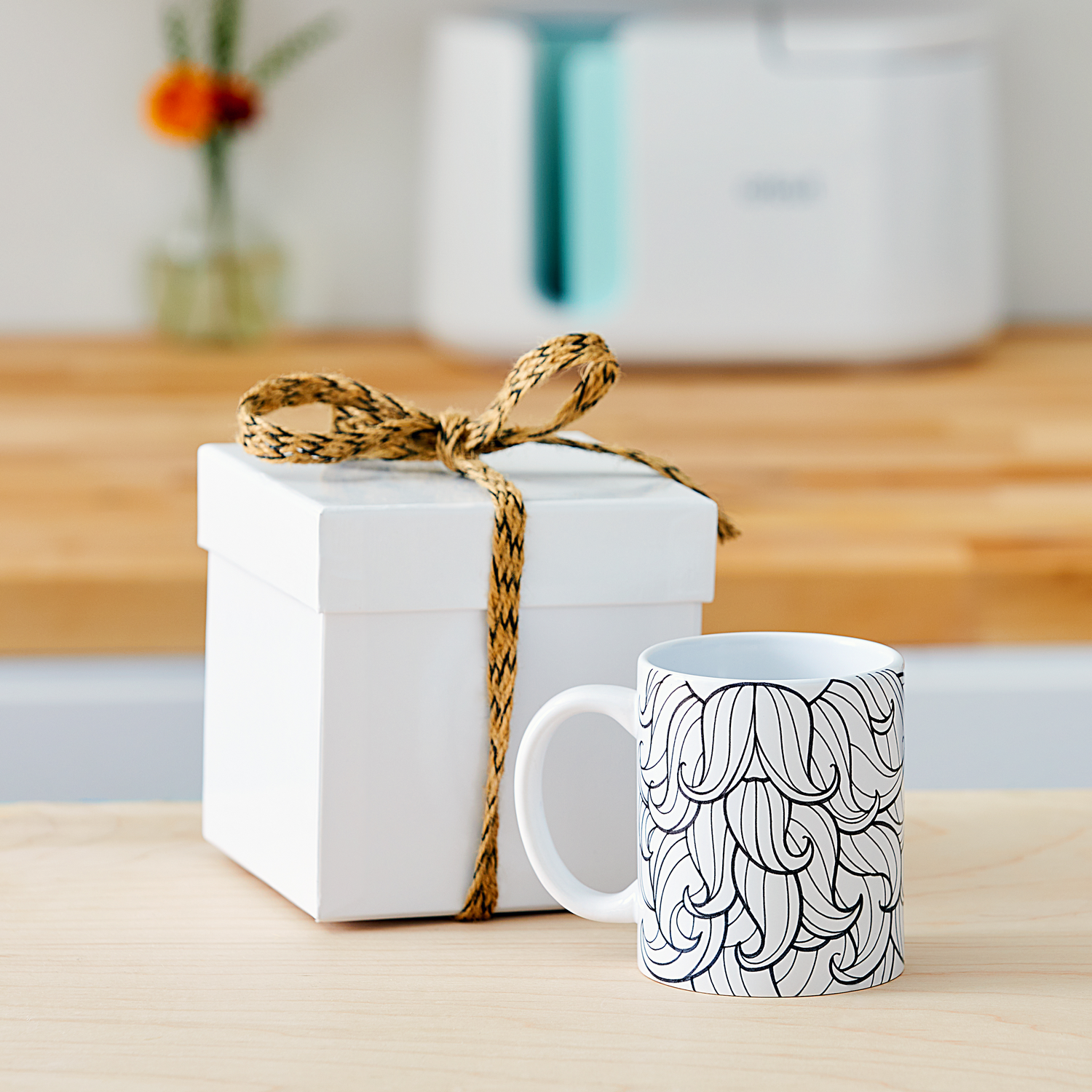 Una caja de regalo blanca atada con cordel junto a una taza blanca con un patrón negro.