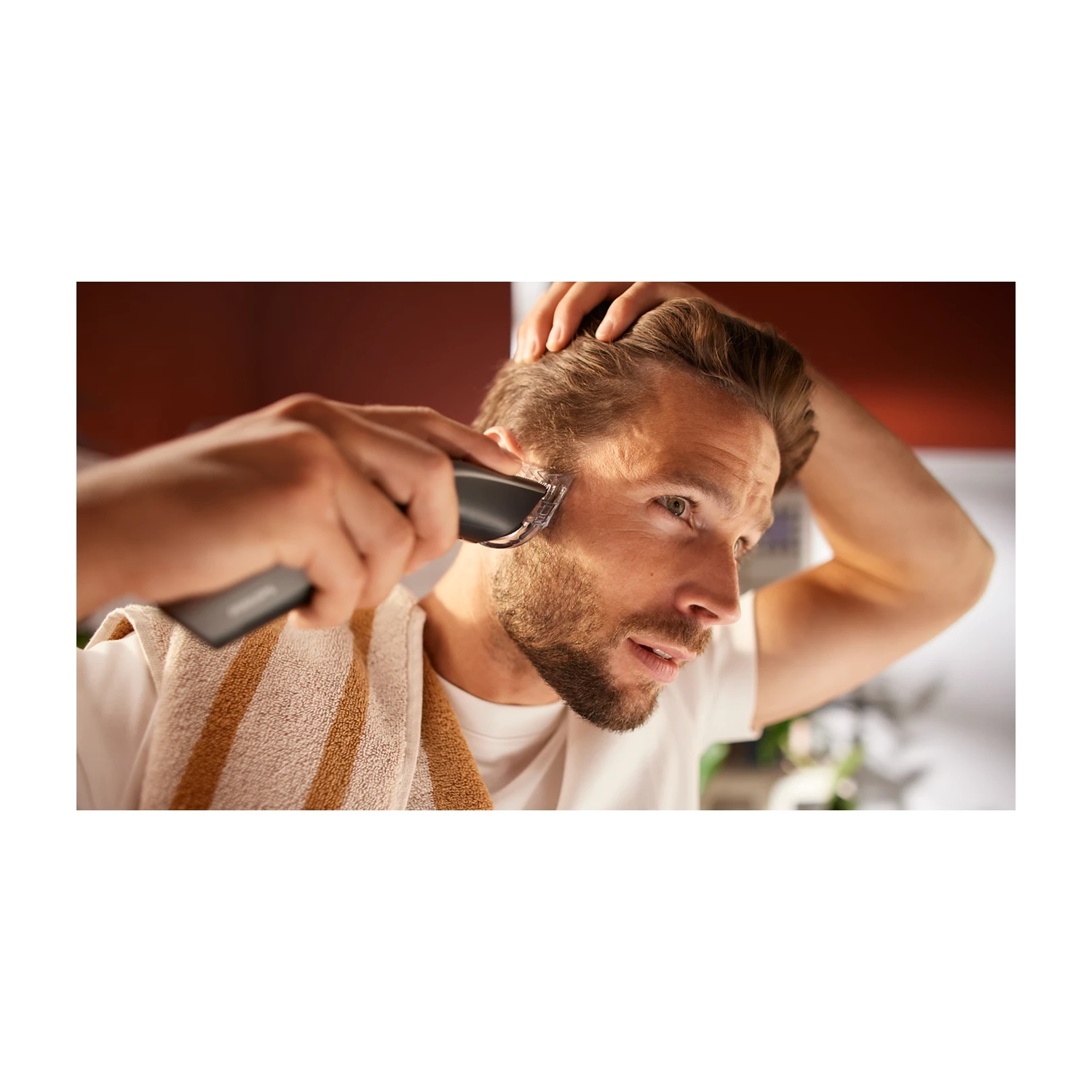 Uomo si taglia barba con rifinitore, mano sulla testa, asciugamano al collo.