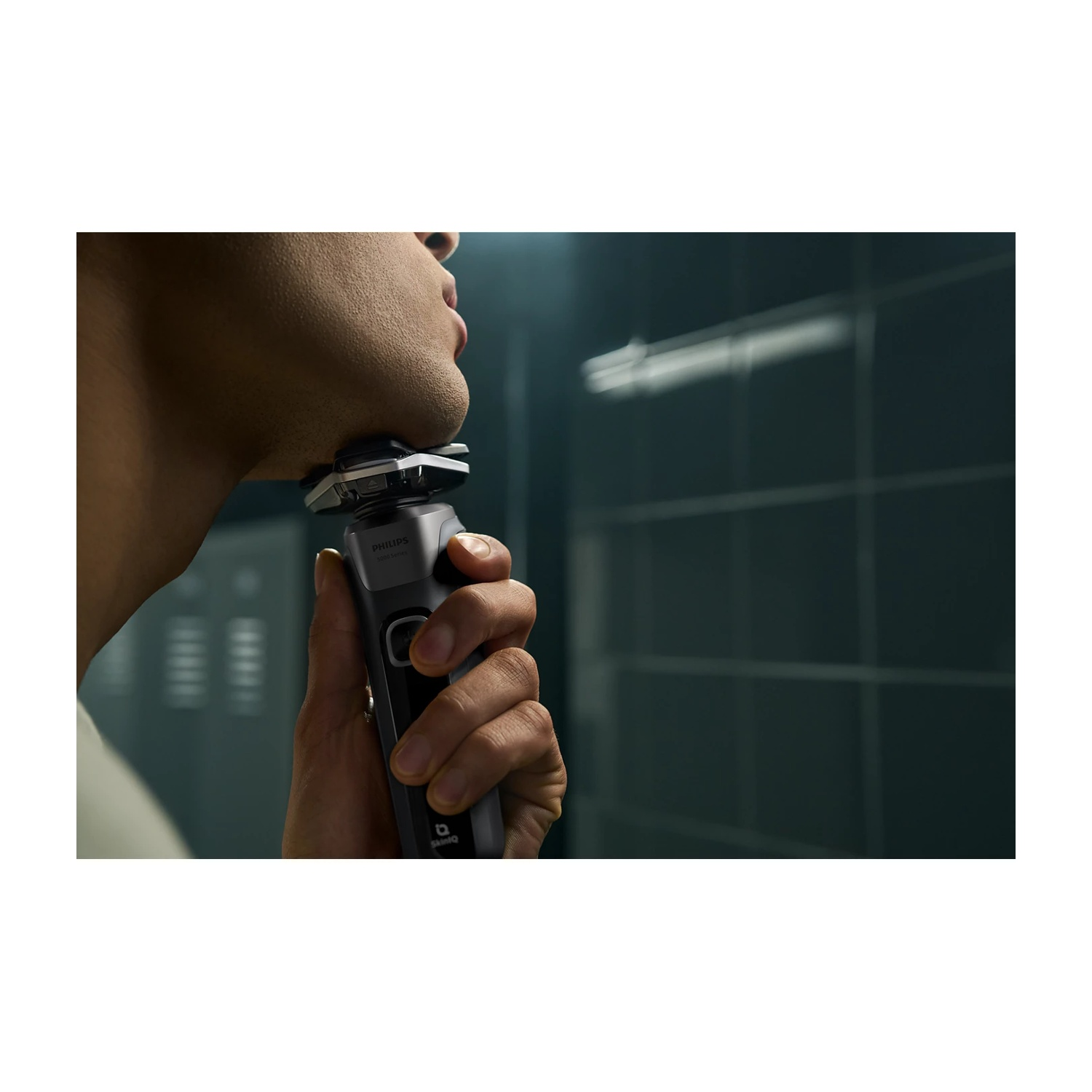 Uomo che si rade con rasoio Philips in bagno. Parete piastrellata scura.