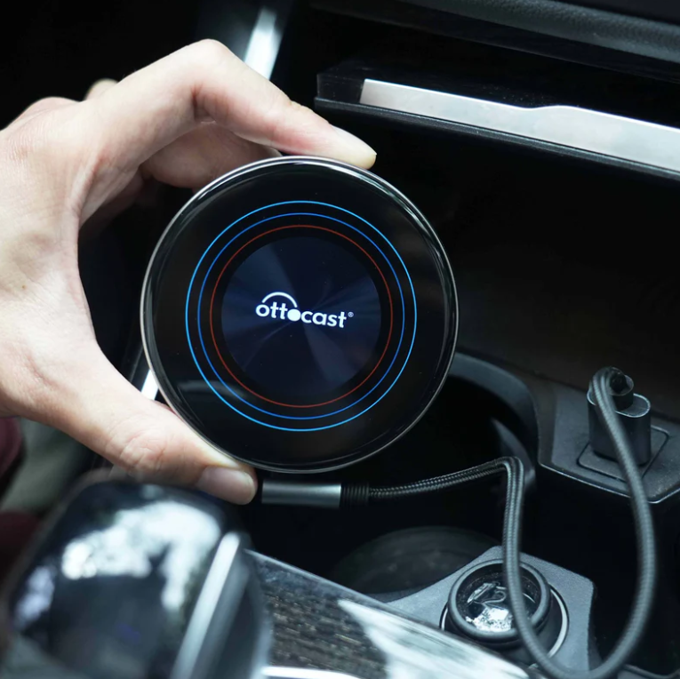 Dłoń trzyma okrągłe urządzenie Ottocast w uchwycie na kubek w samochodzie z kablami.