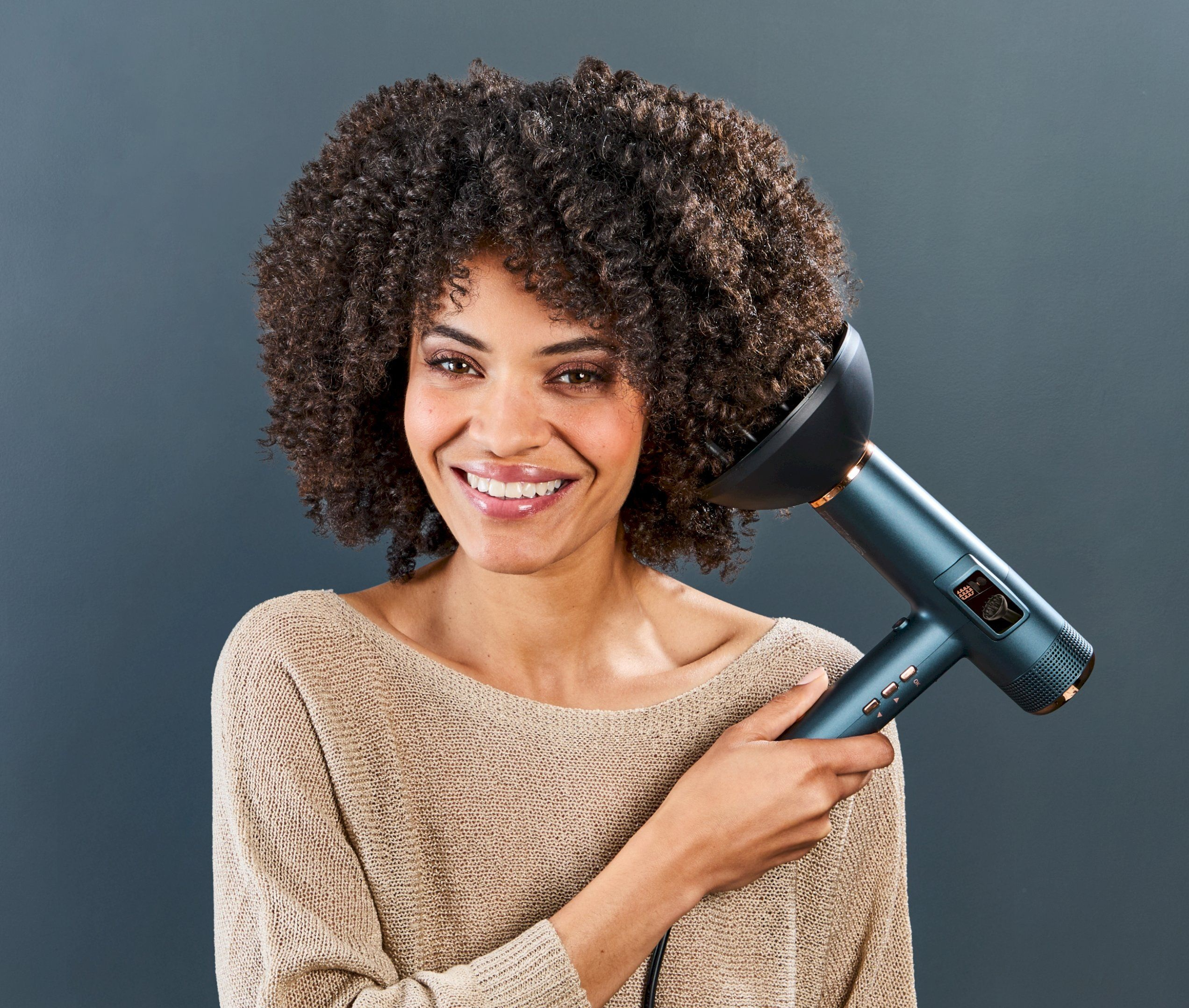 Uśmiechnięta kobieta z kręconymi włosami trzyma turkusową suszarkę do włosów z różowo-złotymi akcentami.