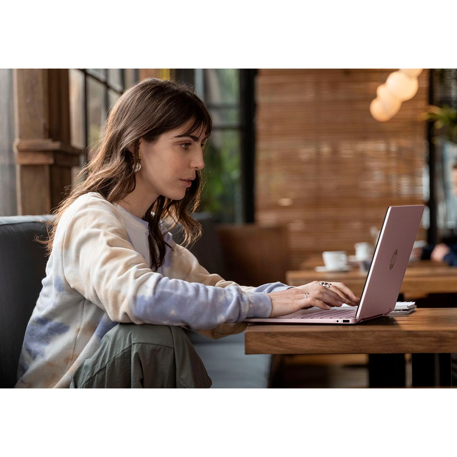 A woman with brown hair types on a pink laptop in a cafe setting.