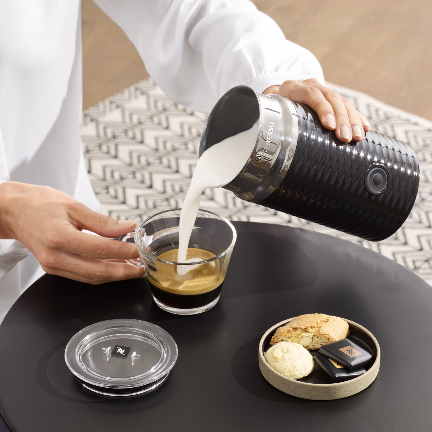 Une personne verse du lait dans un expresso. Des biscuits et des chocolats sont sur la table.