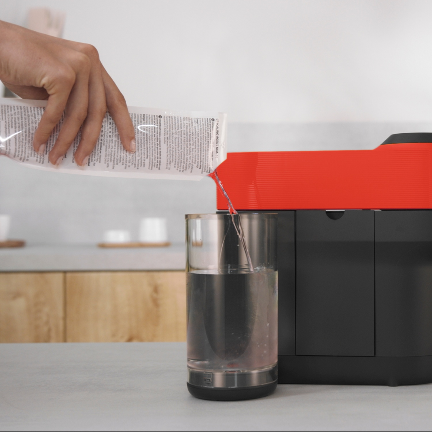 A hand pours liquid from a packet into a glass of water next to a coffee machine.